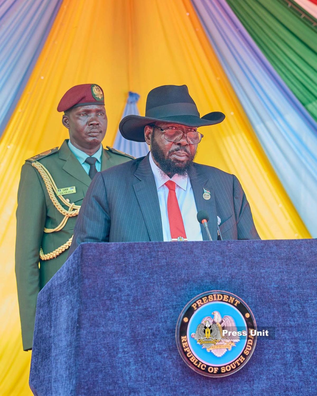 President Salva Kiir Mayardit speaking at a podium during the Juba International Airport Terminal Three inauguration, flanked by a South Sudan People's Defence Forces officer in dress uniform.